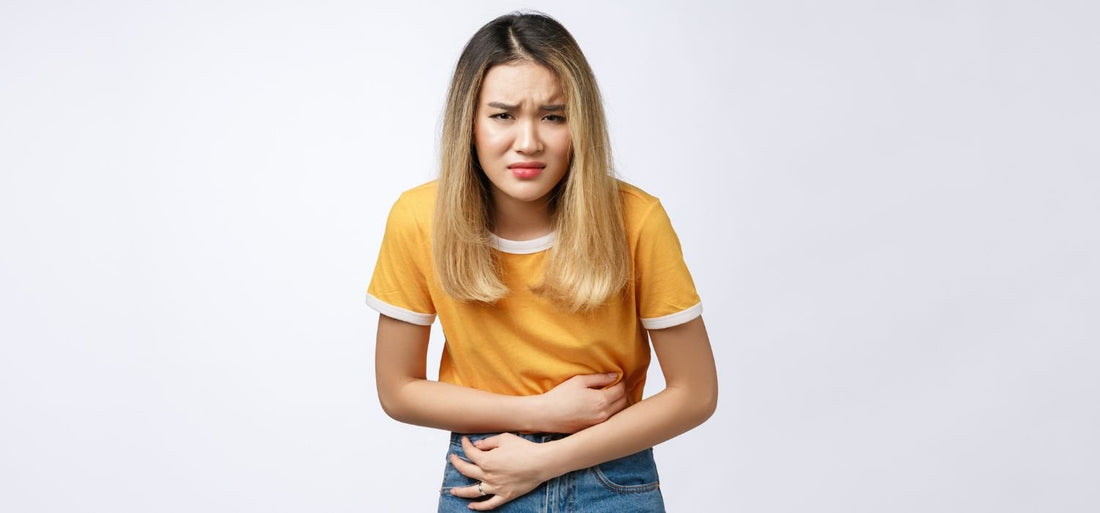 Femme portant un t-shirt jaune et blanc, tenant son estomac avec une expression de douleur ou de malaise, sur un fond blanc uni